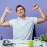 smiling-young-handsome-male-student-sitting-desk-with-school-tools-points-himself-isolated-blue_141793-73127