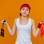 young-fitness-woman-sportswear-with-headband-holding-two-skipping-ropes-looking-confused-shrugging-shoulders-having-doubts-standing-orange-wall_141793-49957